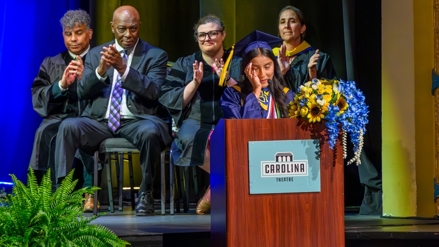 Michelle Amador, who was selected by her classmates to speak, addresses those gathered as Dr. William J. Villalba, world language instructor; Phil Ford, former UNC basketball player and assistant coach; Dr. Mary Margaret Mills-Thomason, director of curriculum and instruction; and Jaclyn Volpe, STEM instructor and community engagement coordinator, look on. 